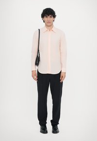 Lichtroze overhemd met lange mouwen en een knoopsluiting, gecombineerd met zwarte broek en zwarte schoenen. Zwarte schoudertas over de schouder.
