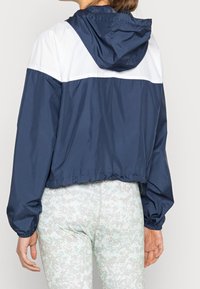 Granatowa krótka kurtka typu windbreaker z białym górnym panelem, wyposażona w kaptur, elastyczne mankiety oraz gładką fakturę.