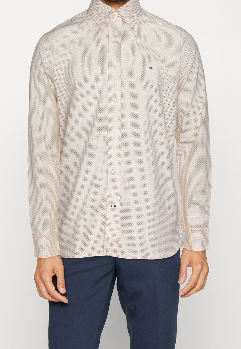 Homme portant une chemise beige à manches longues avec un petit motif à pois et un pantalon bleu foncé, debout devant un fond uni.