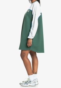 Vihreä ylipitkä mekko, jossa on pitkät harmaat hihat, sinisiä yksityiskohtia ja pieni logo rinnassa. Mallilla on päällään hopeiset tennarit ja valkoiset sukat.