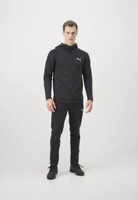 Sudadera negra con capucha y pantalones a juego, hechos de un tejido suave y elástico. La sudadera tiene un cordón y bolsillos laterales; ambos cuentan con un pequeño logotipo.