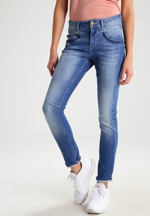 Femme portant un jean slim bleu et des baskets blanches, debout avec une jambe croisée devant l'autre sur un fond uni.
