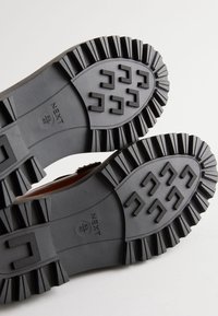 Mustast kummist kinga tallaga, millel on sügavad kulumismustrid, sealhulgas ümarad ja ristkülikukujulised haardepinnad. Märgitud on "NEXT" ja suuruse andmed.