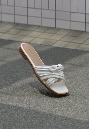 Hvit flat sandal med kryssede remmer og knutedetalj, hvilende på asfalt nær flislagt vegg.