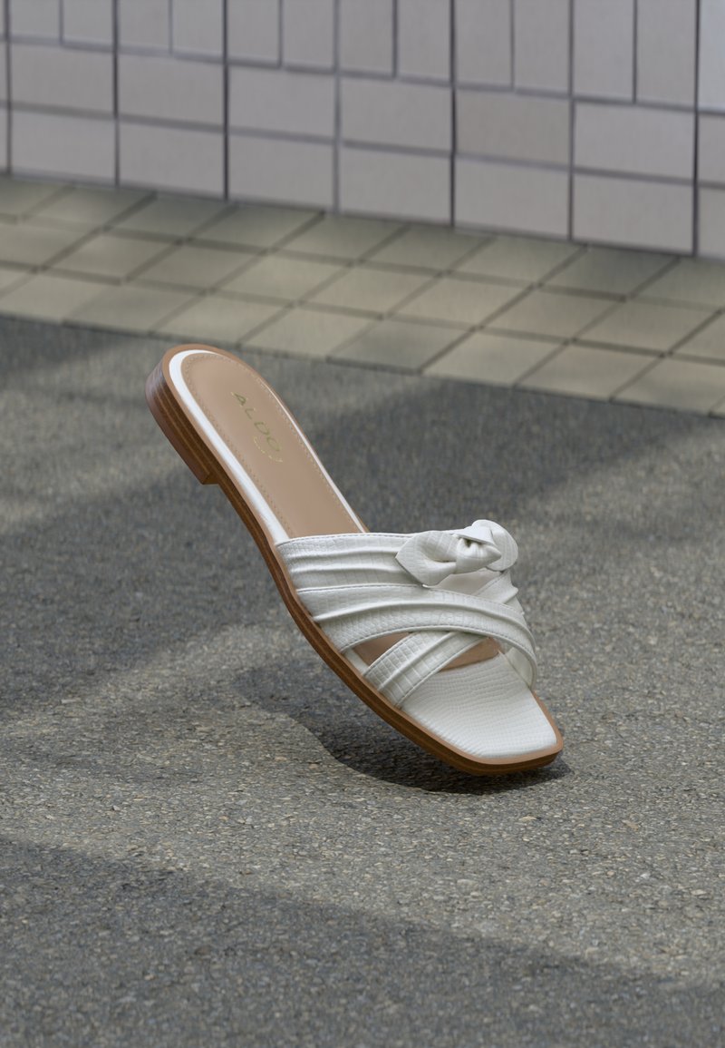 Sandale plate blanche avec des brides croisées et un détail de nœud, posée sur l'asphalte près d'un mur carrelé.