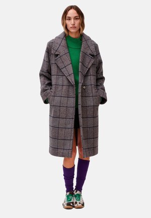 Mujer que lleva un abrigo largo gris de cuadros sobre un suéter verde, calcetines morados hasta la rodilla y zapatillas estampadas con cordones verdes sobre fondo blanco.