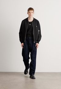 Sort bomberjakke med lynlås, parret med en sort skjorte og løstsiddende mørkeblå bukser. Sorte sko fuldender outfittet.