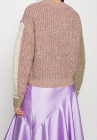 Strickpullover in Rosa mit strukturierten Ärmeln; ein cremefarbener Rippstrickärmel, ein olivgrüner. Getragen über einem glänzenden lila Satinrock.
