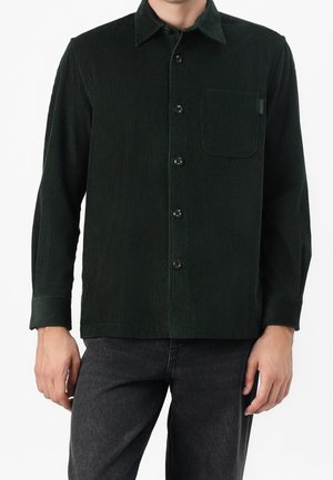 Mann trägt dunkelgrünes Cordhemd mit Brusttasche und schwarze Jeans vor weißem Hintergrund.