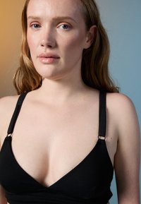 Γυναίκα με ανοιχτόχρωμο δέρμα και μακριά ανοιχτό καστανά μαλλιά φοράει ένα μαύρο σουτιέν, στέκεται μπροστά σε ένα δίχρωμο φόντο με κίτρινα και μπλε τόνους.