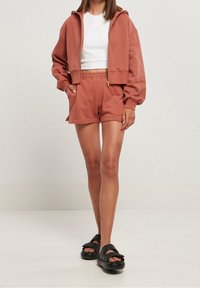 Felpa corta in terracotta con cerniera, abbinata a pantaloncini coordinati. Il tessuto sembra morbido; i pantaloncini hanno tasche laterali e una vestibilità comoda.