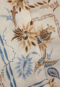 Chemise à boutons avec un fond crème, ornée de motifs floraux bleus et marron, boutons ronds et tissu texturé.