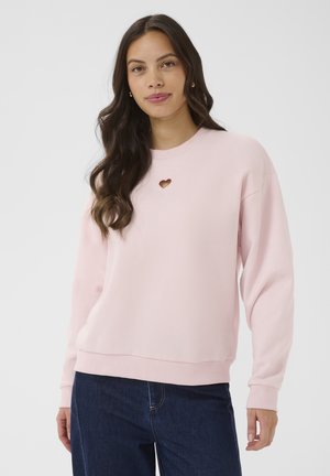 Žena s dlhou tmavou vlasmi, oblečená v svetloružovej mikine s malým výrezom v tvare srdca na hrudi a tmavomodré rifle.