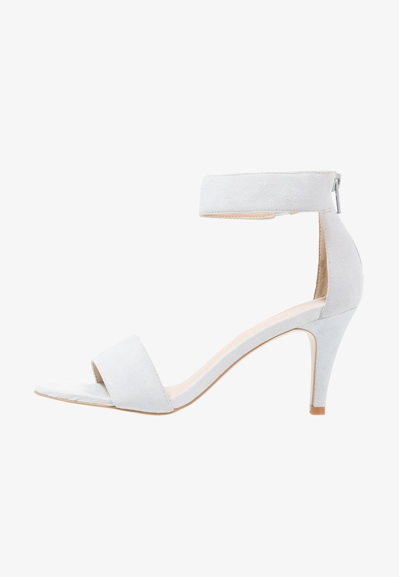 Sandale blanche en daim à talon haut, bout ouvert, bride à la cheville et fermeture éclair au dos.