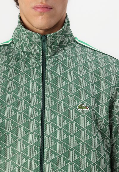 Veste zippée verte avec un motif géométrique, col haut et logo discret. Comprend des rayures contrastantes blanches et vertes sur les épaules.