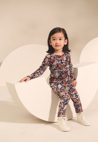 Marinblå långärmad topp och leggings med blommönster i stretchigt tyg, båda bärs med vita höga sneakers, sittande på en skulptural vit form.