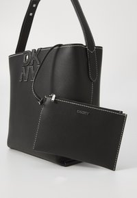 Melnas ādas iepirkumu soma ar kontrastējošu baltu šuvju, ar iegrieztu "DKNY" logotipu un noņemamu rāvējslēdzēja kabatu.