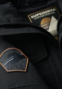 Sort jakke med et broderet Superdry-patch i orange og sort, med lynlåslukning og et guld-accentueret mærkelabel.