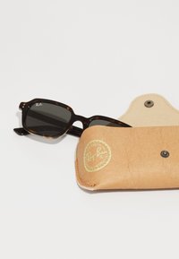 Ochelari de soare tortoise maro lângă o carcasă din piele maro deschis cu un logo circular aurit și închidere tip snap.