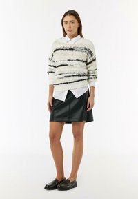 Un pull blanc duveteux avec des rayures noires, superposé sur une chemise blanche à col. Jupe en cuir noire en forme de A et mocassins noirs.