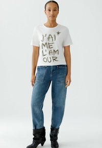 Weißes Baumwoll-T-Shirt mit schwarzem Text "J'AI ME L'AM OUR" in handgezeichneter Schrift. Kombiniert mit hochgeschnittenen blauen Jeans und schwarzen Stiefeln.