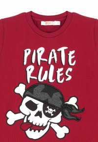 Rød bomulds-T-shirt med et sort-hvidt piratskull grafik med krydsede knogler og teksten "PIRATREGEL" i fed hvid skrift.