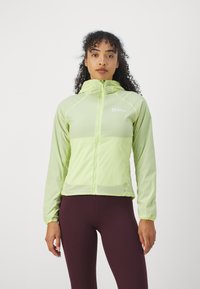 Giacca a zip verde chiaro con cappuccio, realizzata in un materiale morbido e leggero, con maniche raglan. Include il logo sul lato superiore sinistro.