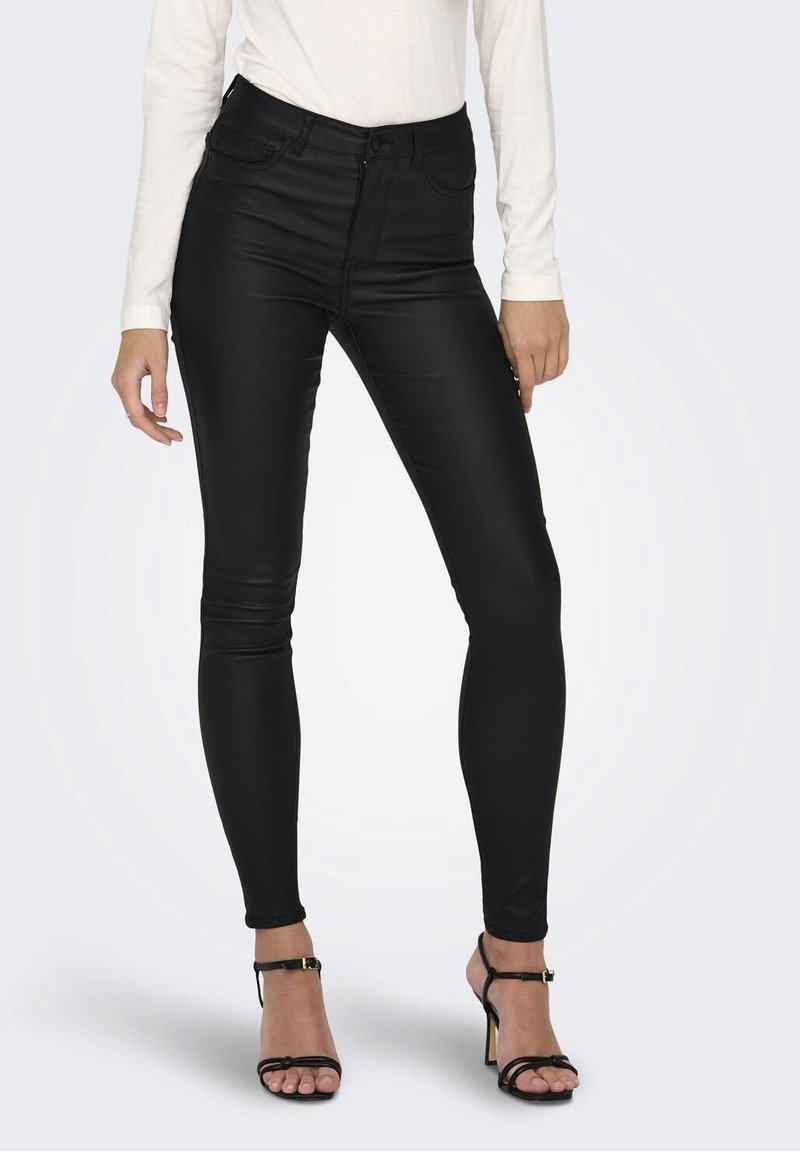 Femme portant un pantalon skinny noir enduit taille haute avec poches avant et des sandales noires à talons hauts, debout sur fond blanc.
