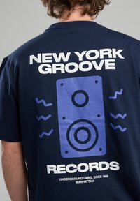 Navy blue t-shirt featuring a large graphic of a speaker in purple and white text stating “NEW YORK GROOVE RECORDS” on the back.