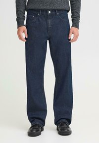 Pantalones de mezclilla azul oscuro con un diseño de pierna recta, que cuentan con cinco bolsillos y herrajes plateados. Llevados con zapatos de cuero negros.