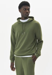 Sudadera verde oliva de corte holgado, con capucha ajustable y textura sutil. Combinada con pantalones verde oliva a juego.