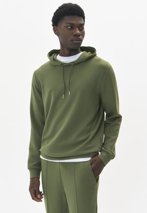 Olijfgroene hoodie met een ontspannen pasvorm, voorzien van een trekkoordenkap en een subtiele textuur. Gecombineerd met bijpassende olijfgroene broek.