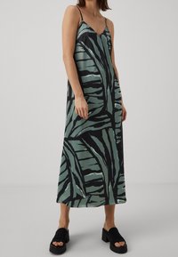 Vestido maxi sin mangas en estampado de hojas de palma en verde y negro, tela suave, escote en V, con tirantes delgados ajustables. Lleva sandalias negras de tacón cuadrado.