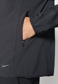 Sort Nike jakke med en lynlåslomme og elastiske manchetter. Materialet ser glat ud med en mat finish. Diskret logo detalje inkluderet.