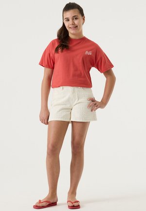 Camiseta roja de algodón con mangas cortas, combinada con pantalones cortos blancos con textura. El modelo también lleva chanclas rojas. Fondo liso.