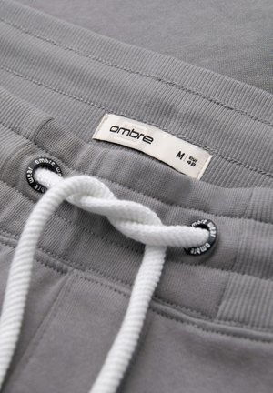 Gros plan sur la ceinture d'un pantalon de survêtement gris avec un cordon blanc, des œillets métalliques et une étiquette en tissu "ombre" taille M (EUR 48).