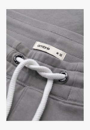 Close-up van de tailleband van een grijze joggingbroek met witte trekkoorden, metalen oogjes en een stoffen label met "ombre" maat M (EUR 48).
