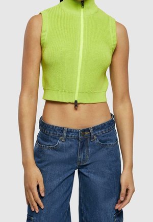 Bezrekawnikowy zielony neonowy krótki sweter z przodu na zamek, noszony z wysokim stanem niebieskich dżinsów na osobie stojącej.