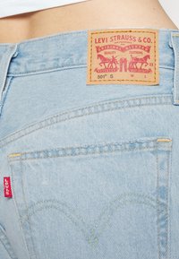 Light blue denim jeans feature a classic back patch with red and yellow branding, gold stitching, and a curved pocket design.