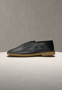 Chaussure noire en cuir à enfiler avec une semelle en caoutchouc beige, présentant un design plié et un talon bas, exposée sur une surface claire.