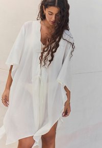 Balts, viegls kaftans ar brīvu, gaisīgu dizainu, V veida kakla izgriezumu un regulējamu jostu. Iespēj wide piedurknes un sānu izgriezums, kas nodrošina kustību.