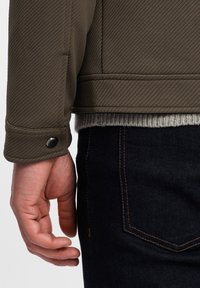 Olivgrön quiltad jacka med en texturerad yta, tryckknappslås vid muddarna och en grå ribbstickad tröja under, kombinerad med mörk denim.