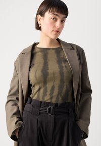 Maslinasto zeleni rebrasti top ispod sive blejzera, uparen s crnim hlačama visokog struka koje imaju okruglu kopču. Vidljive su glatke teksture.
