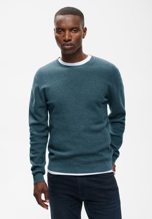 Homme portant un pull à manches longues bleu sarcelle par-dessus une chemise blanche et un pantalon sombre, debout devant un fond clair uni, les mains le long du corps.