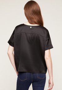 Top de satén negro con mangas cortas, que presenta un escote redondeado y un pequeño detalle de botón en la parte posterior, mostrado con jeans azules.