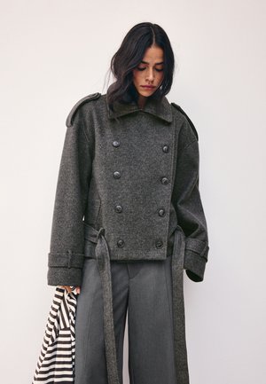Femme aux cheveux foncés portant une veste croisée gris foncé et un pantalon gris, tenant un sac en tissu rayé noir et blanc.