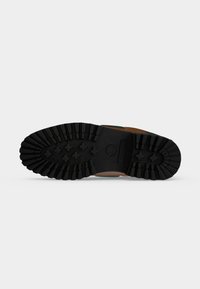 Semelle d'une botte marron et verte avec des motifs épais en caoutchouc noir pour la traction, présentée sur un fond gris clair uni.