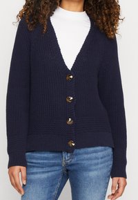 Cardigan lavorato a maglia blu navy con scollo a V, sette bottoni dorati sul davanti e polsini e orlo a coste. Tessuto con motivo testurizzato in tutto il capo.