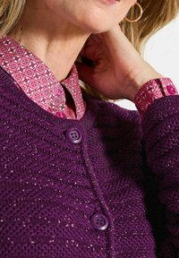 Lila gestrickte Strickjacke mit strukturierter Oberfläche, ausgestattet mit zwei lila Knöpfen. Darunter sorgt ein rosa gemustertes Hemd mit Kragen für einen Kontrast.