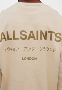 Μπεζ φούτερ με έντονα καφέ γράμματα: «ALLSAINTS», ιαπωνικούς χαρακτήρες και «LONDON» στο κέντρο. Μαλακή υφή και άνετη εφαρμογή.